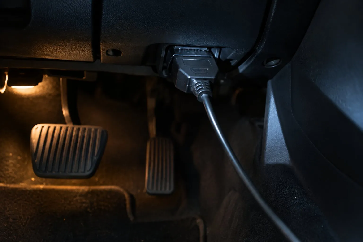 Fußraum eines Autos im Halbdunkel: Unter dem Armaturenbrett steckt ein neutraler schwarzer Stecker mit Kabel in der Diagnosebuchse, darunter Brems- und Gaspedal (KI-generiertes Symbolbild)